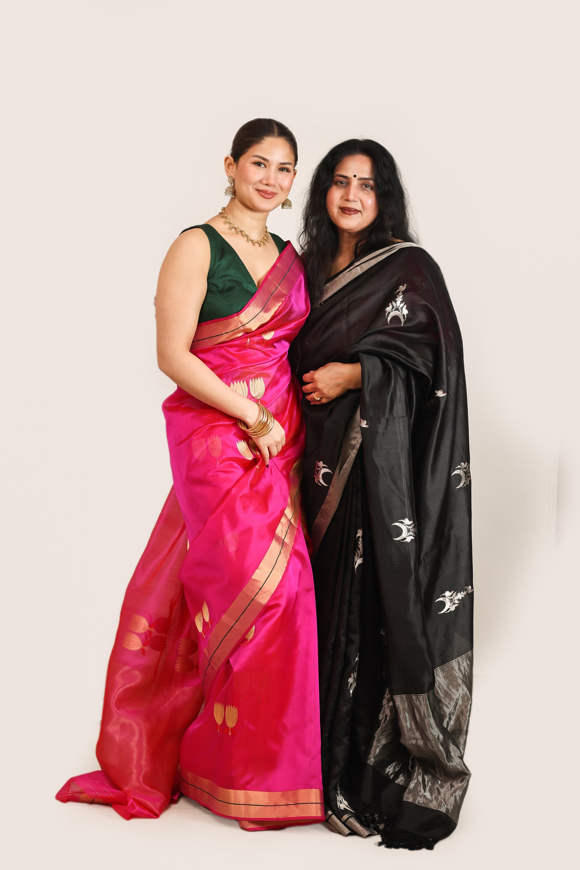 Two women in sarees standing side by side against a plain background